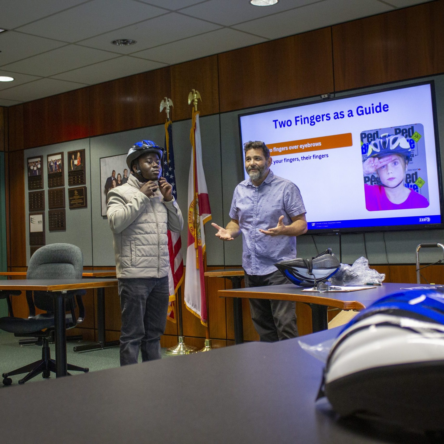 The male trainer demonstrates a helmet fitting on a male student at the front of the room.