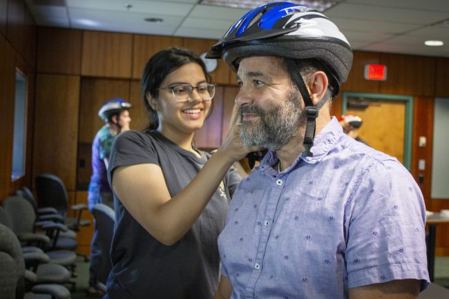 Helmet Fitting Female student practices fitting a bicycle helmet on an adult male. Her hands are adjusting the helmet straps of the helmet on his head.