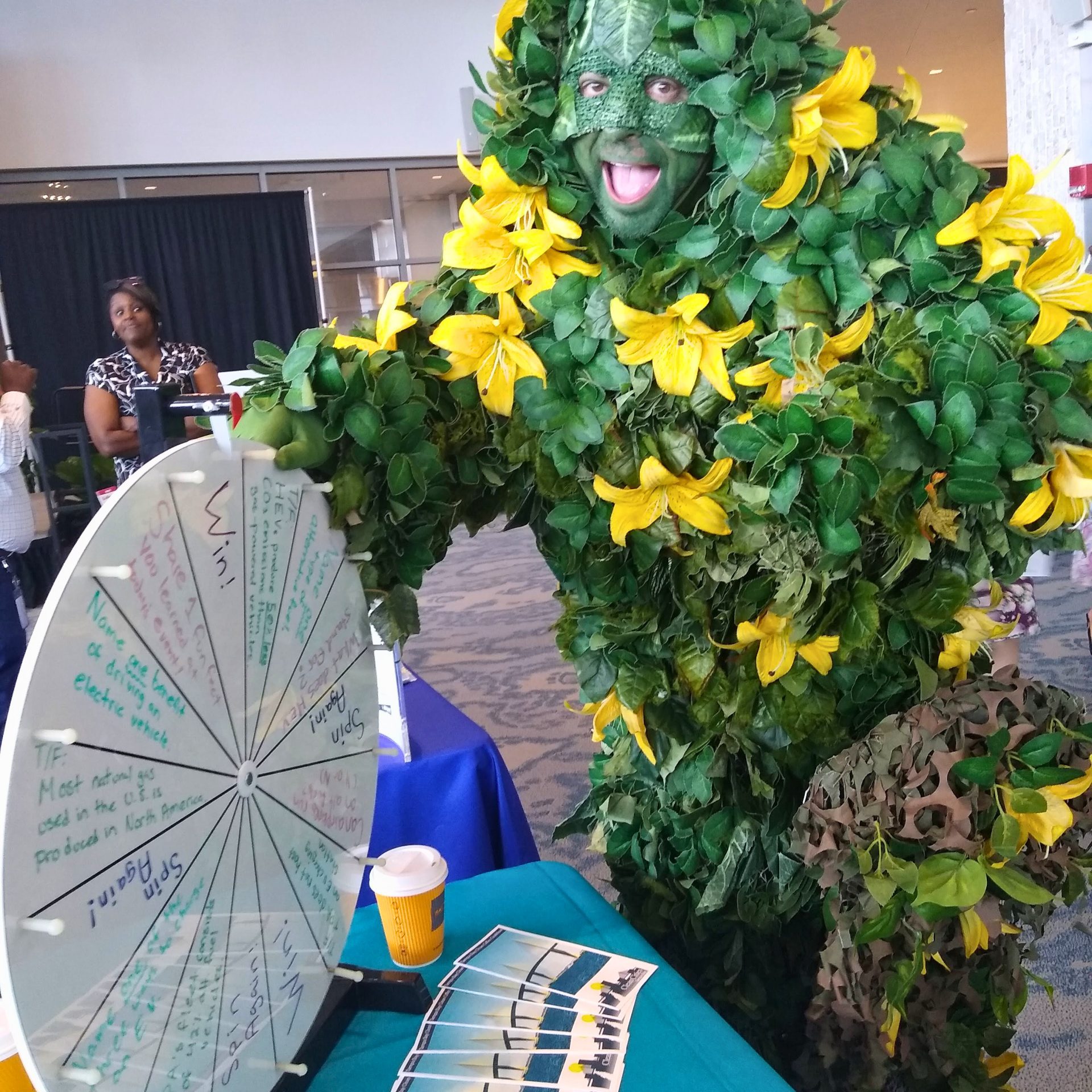 Person dressed in plant costume stands next to exhibitor table with roulette wheel.