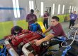 Two male mechanics work on a bus engine while others watch.