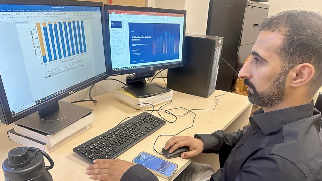 Adult male sits in from of a computer with two monitors, looking at data graphs.
