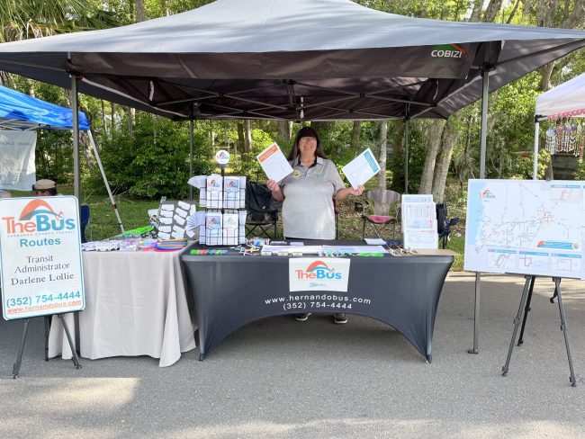 thebus-exhibit1 Outdoor community event. woman stands behind exhibitor table covered in informational materials and give-aways, holding flyers in her hands.