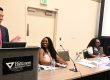 presentation with male standing at podium smiling and three women sitting at presenter table in front of room.