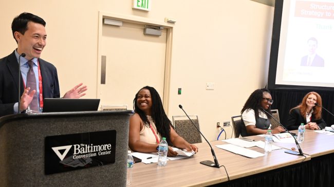 0707-Rail-Workforce-1792×1002-1 presentation with male standing at podium smiling and three women sitting at presenter table in front of room.