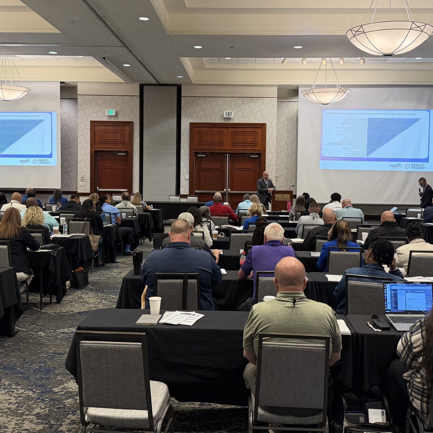 large meeting room with people sitting at desks, facing front of room watching a training presentation