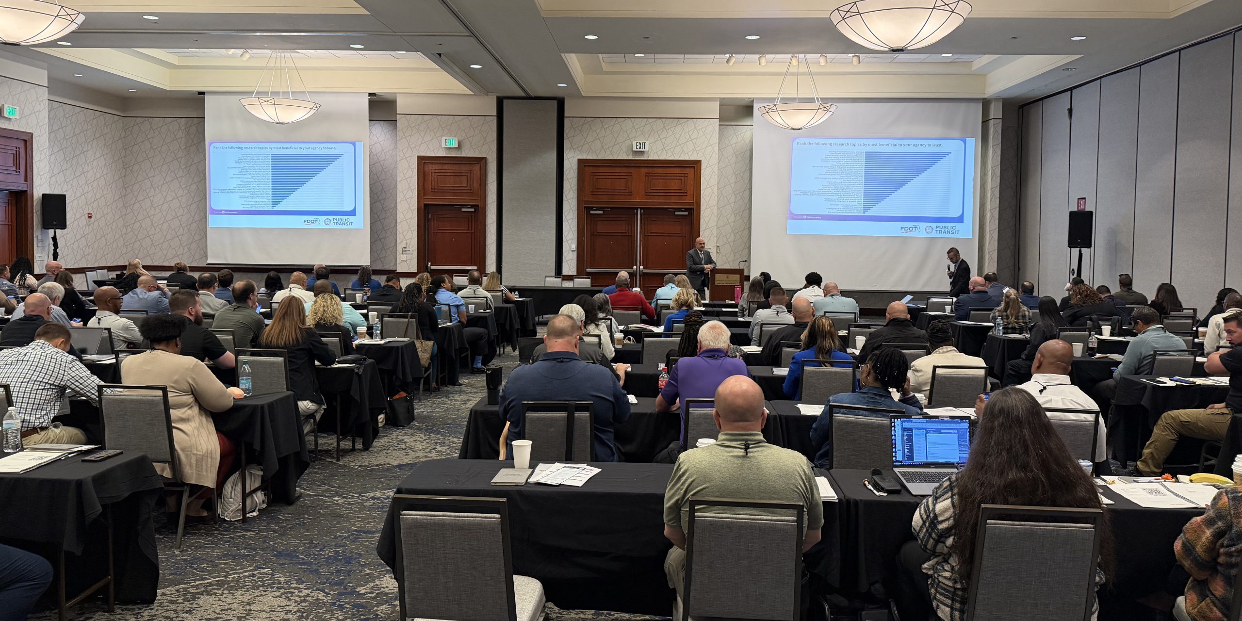 large meeting room with people sitting at desks, facing front of room watching a training presentation