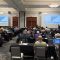 large meeting room with people sitting at desks, facing front of room watching a training presentation