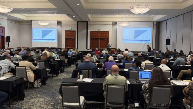 large meeting room with people sitting at desks, facing front of room watching a training presentation