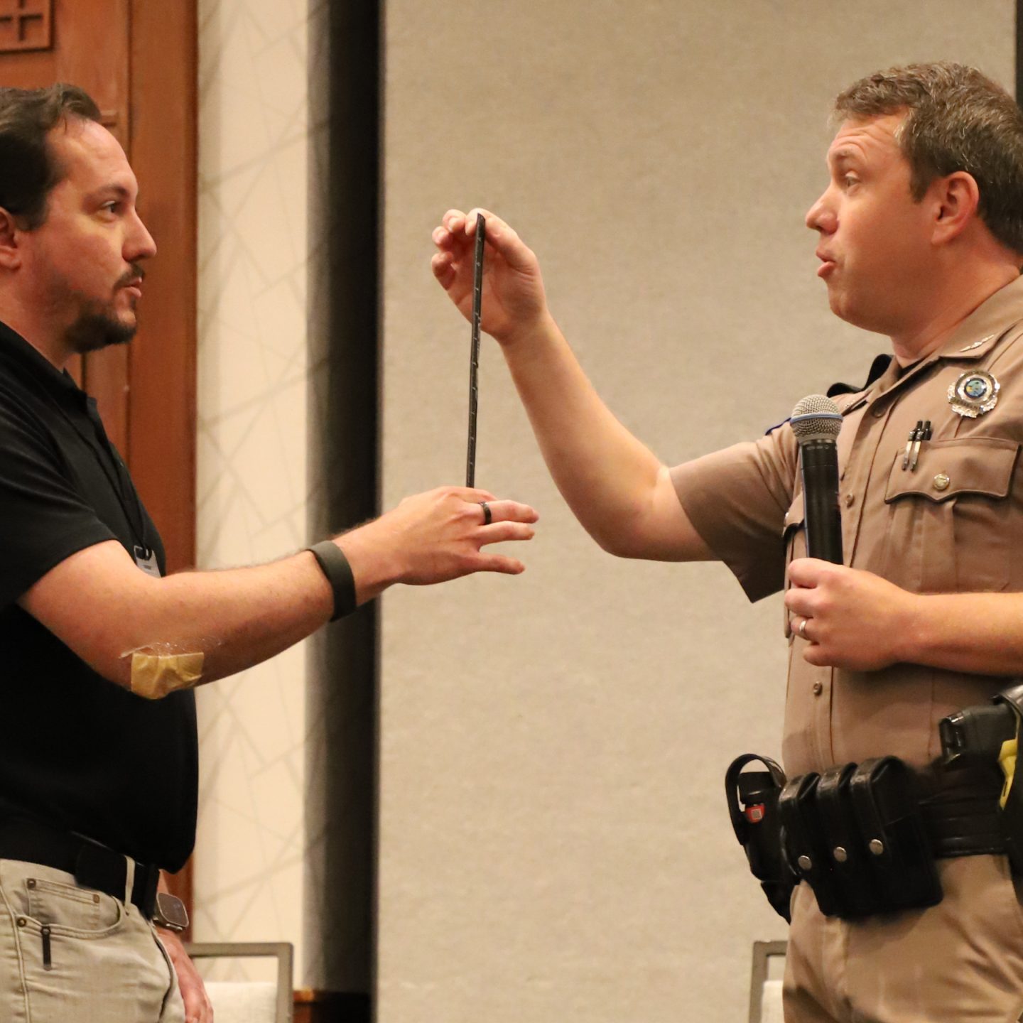 two men in a meeting room face each other holding a stick together. one man holds it from the bottom end. the other man, a law enforcement officer, holds it from the top end.