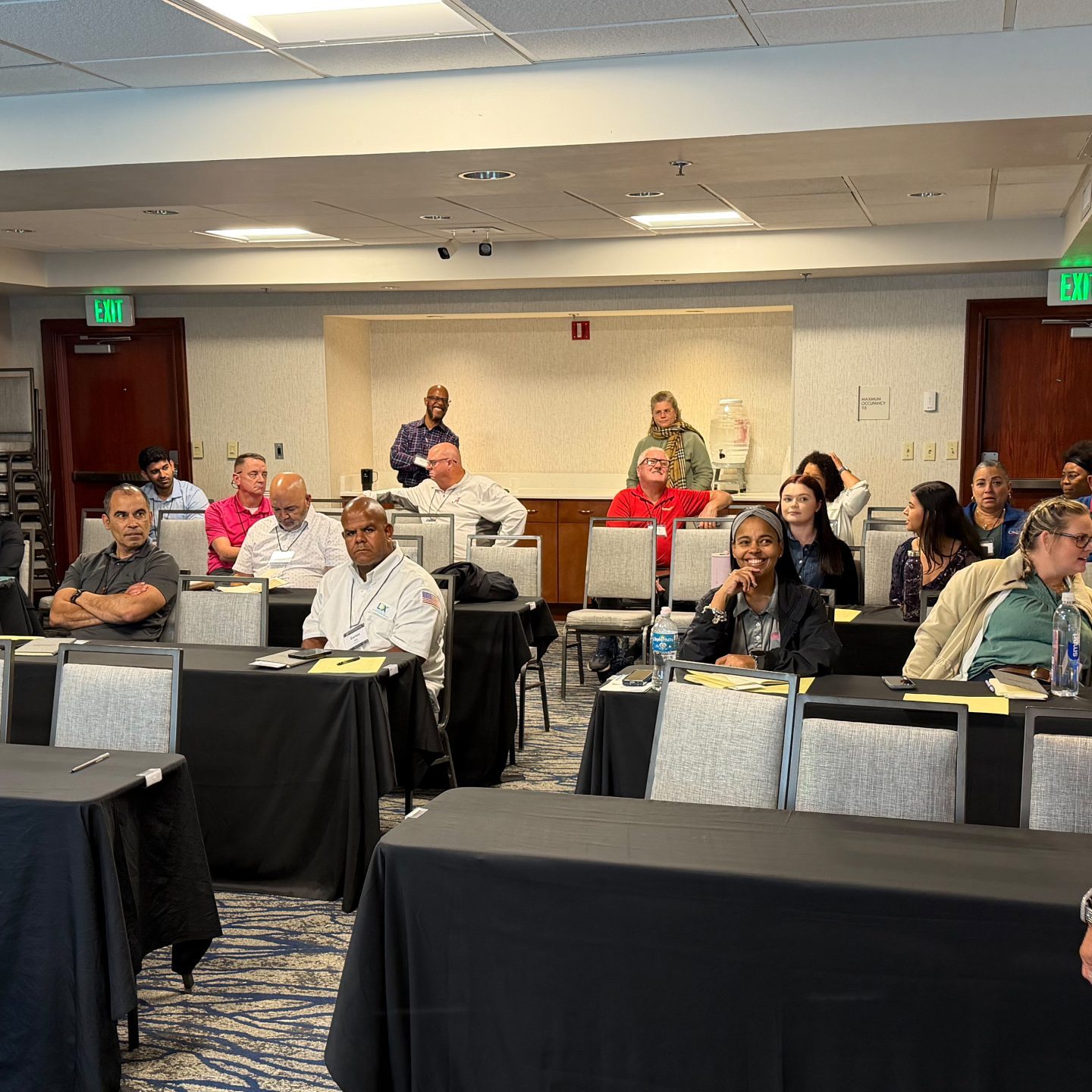 Presenter at front of room addresses seated audience in training room.