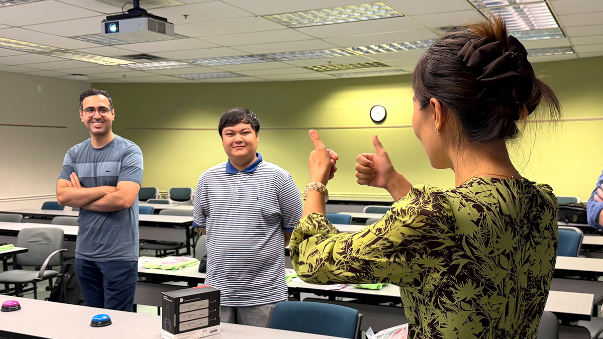 Presenter gives two thumbs up to two competitors standing in front of buttons during quiz game.