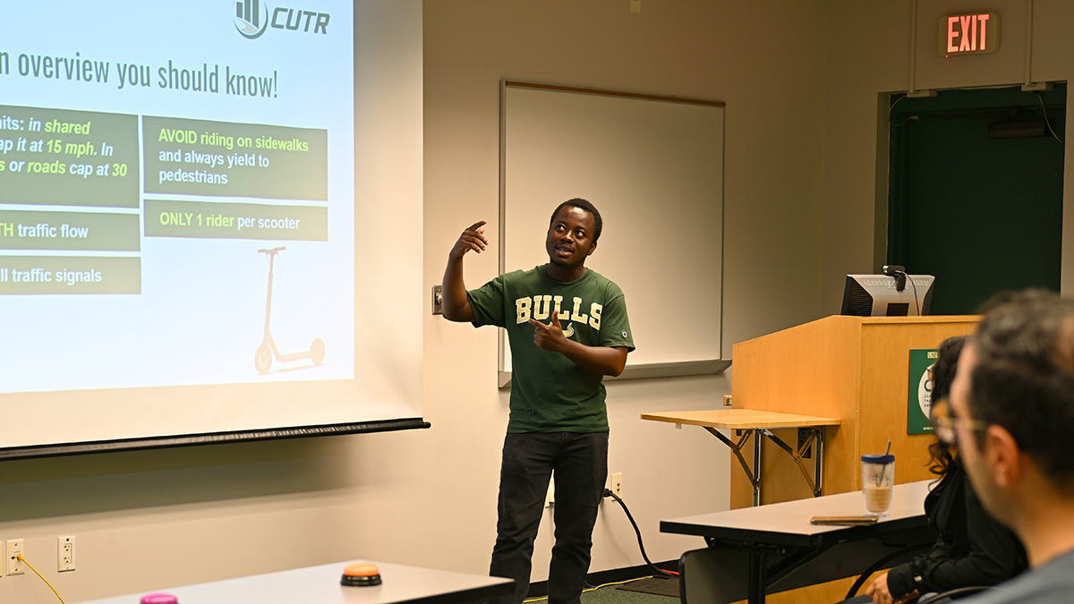 young man stands next to slide projection, giving presentation to group