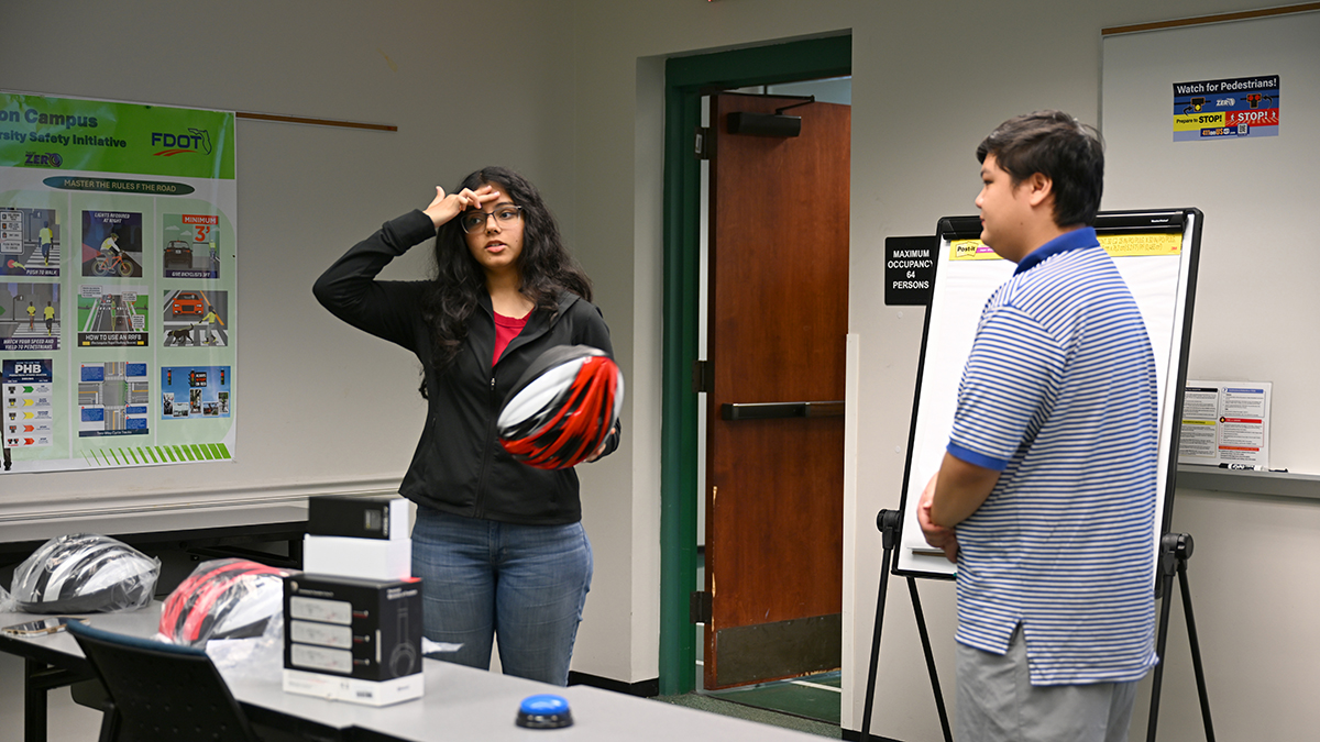 young man and woman stand in front of room. woman holds a bicycle helmet in her hand while using her other hand to touch her forehead, noting helmet placement.