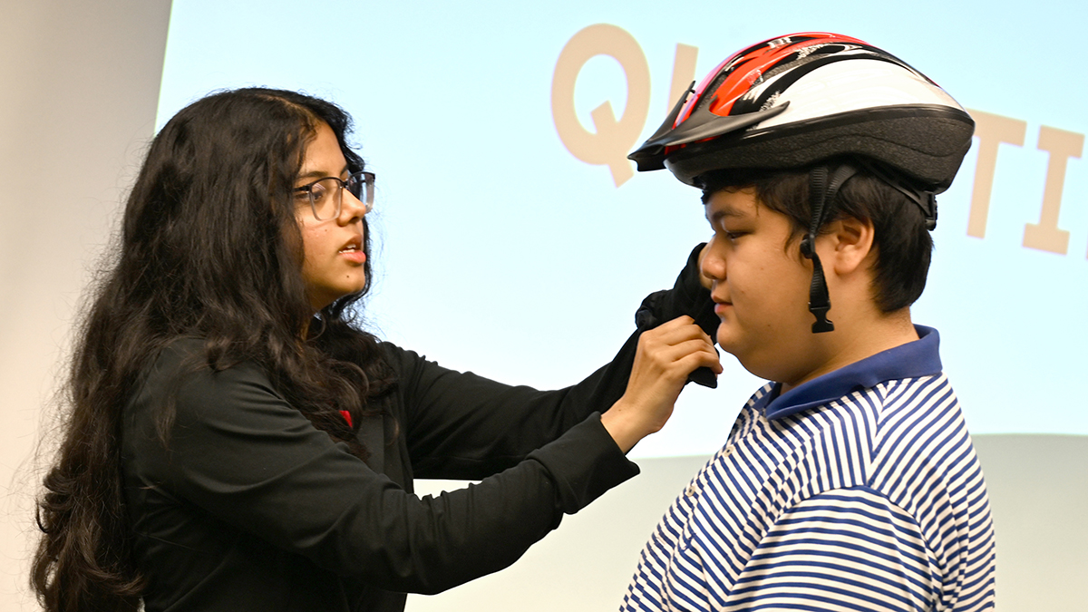 young woman demonstrates how to put on a bicycle helmet on a young male volunteer