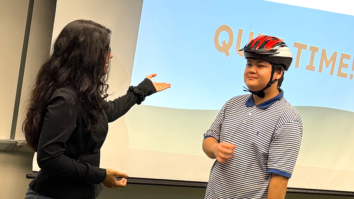 young man and woman stand in front of room. woman gestures toward man wearing bicycle helmet
